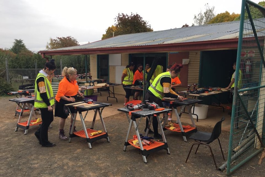 Women and girls in high visibility vests and shirts working at tool benches in front of a shed