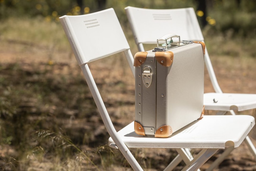 A silver and tan briefcase sits on a white chair outside on the grass.