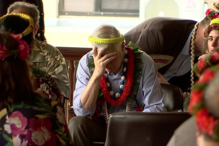 A man in business attire and ceremonial garlands sits with his hand over his face, looking unwell.