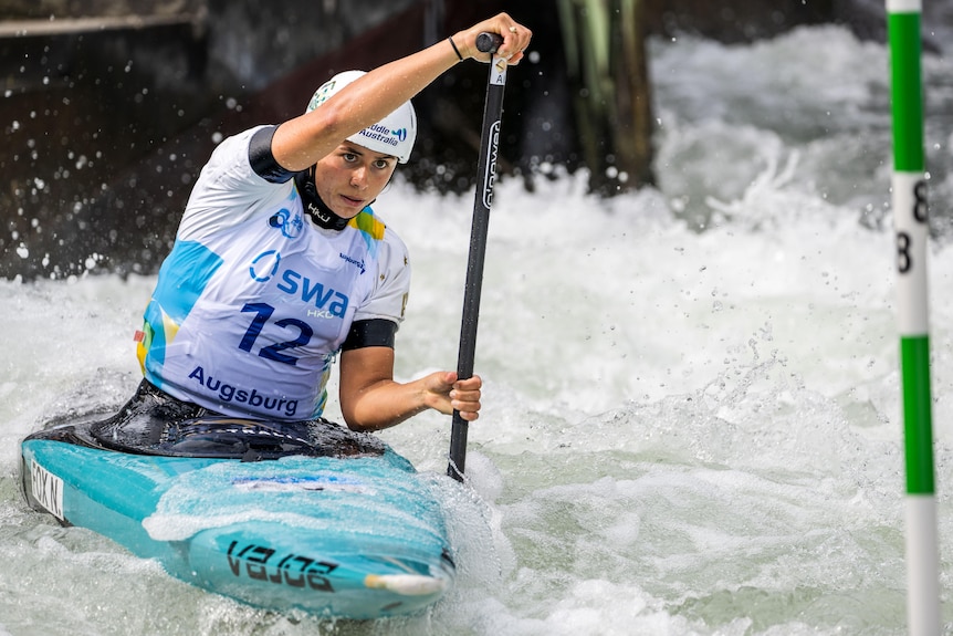 Noemie Fox competes in the women's canoe heats run. She is pictured with a blue canoe and a top with Augsburg on the bottom