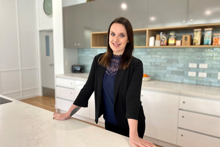 Woman in corporate attire stands in a large kitchen with white marble bench.