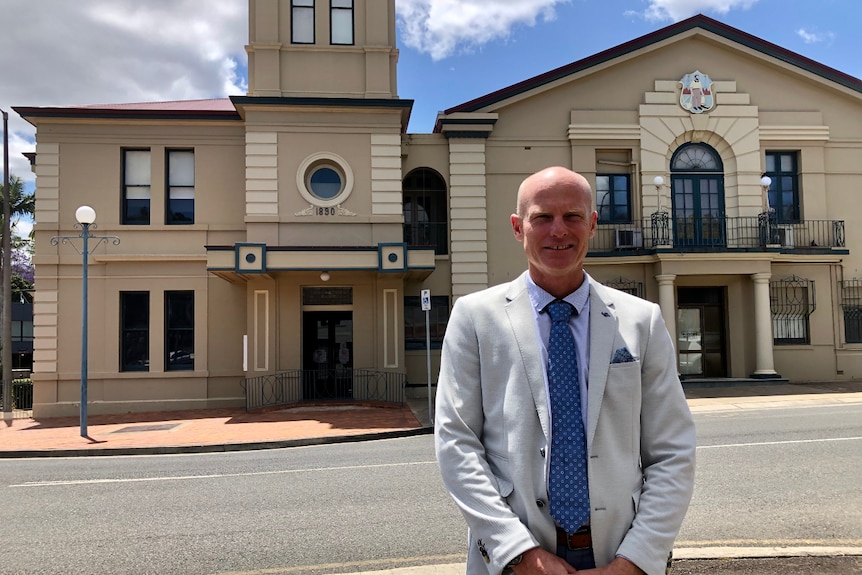 A bald man wearing a suit standing in front of council chambers.