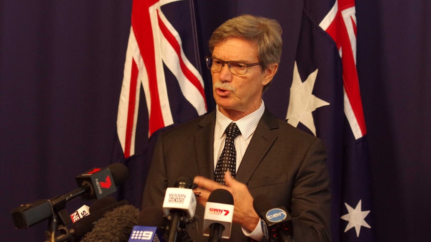 Mike Nahan speaking in front of microphones with Australian flags in the background.