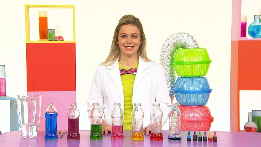 Rachael on the Play School Science Time set wearing a lab coat with plastic bottles filled with coloured water