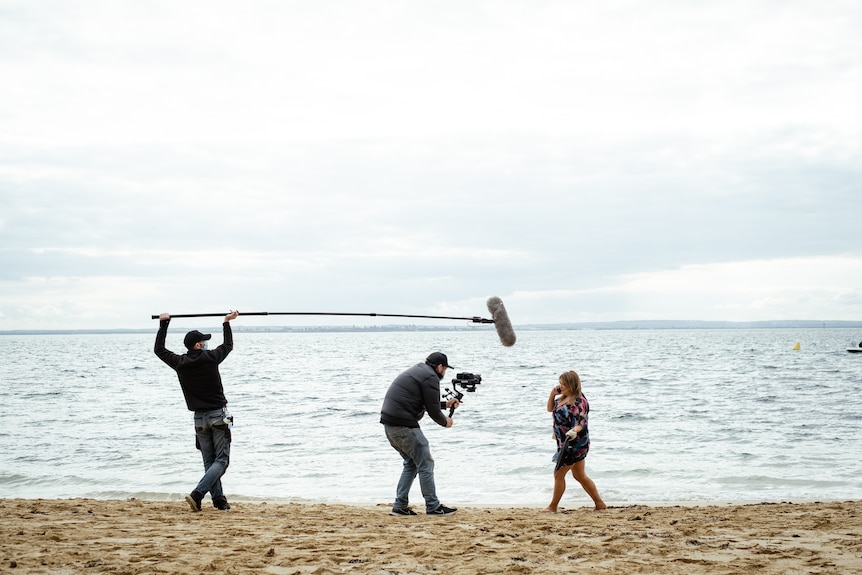 A camera operator, a sound technician and an actor filming a scene of Neighbours on a beach.