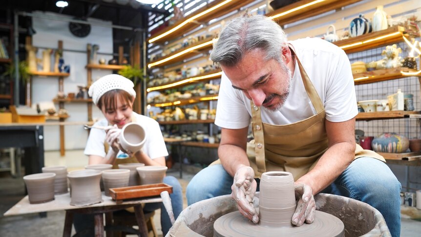 In a light room filled with shelves of ceramics, a smiling woman paints a vase and smiling man molds clay on wheel.
