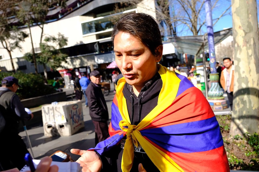 A man wearing a Tibetan flag on his back holds his hands open as he speaks to a reporter.