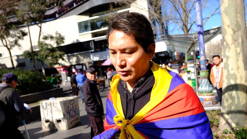 A man wearing a Tibetan flag on his back holds his hands open as he speaks to a reporter.