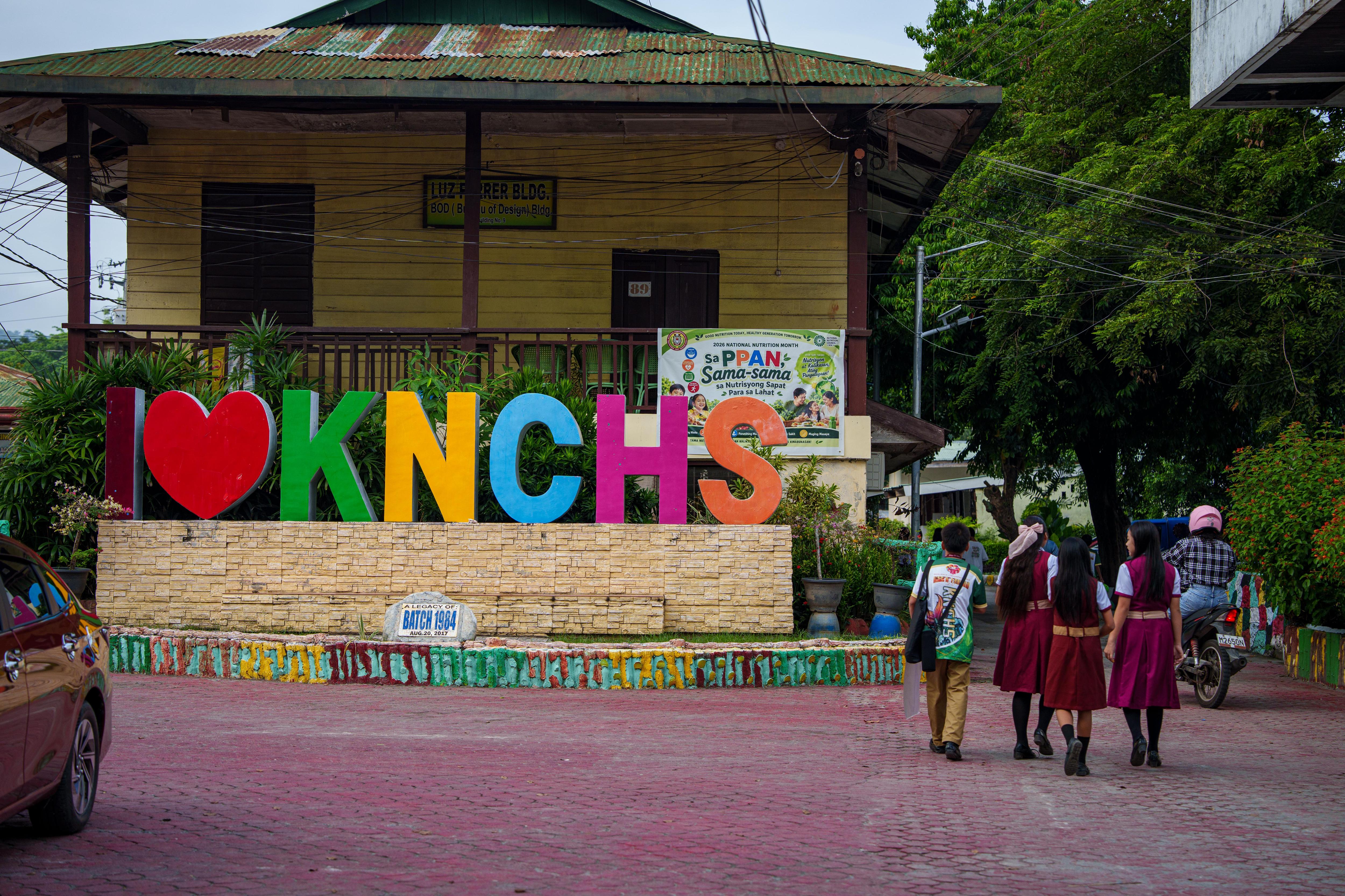 School students walk past a colourful sign outside a building that says "I (love heart) KNCHS".