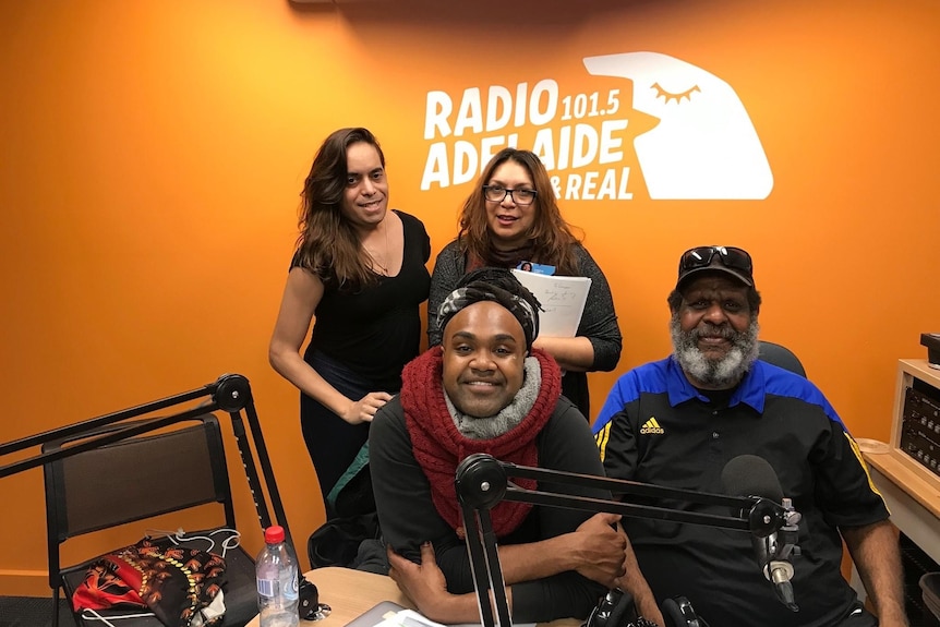 A group of people in an orange radio studio smile against half-hidden sign saying Radio 101.5 Adelaide real.