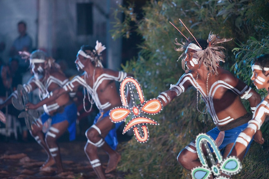 Indigenous dancers perform wearing traditional body paint and costumes, including headdresses made of feathers and gum leaves.