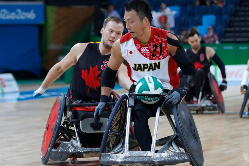A Japanese wheelchair rugby player holds the ball in his lap as he protects it from the Canadian player behind him.