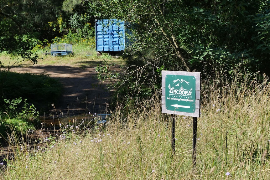 A sign with the Arcoora logo and an arrow pointing to a dirt driveway which runs through a creek.