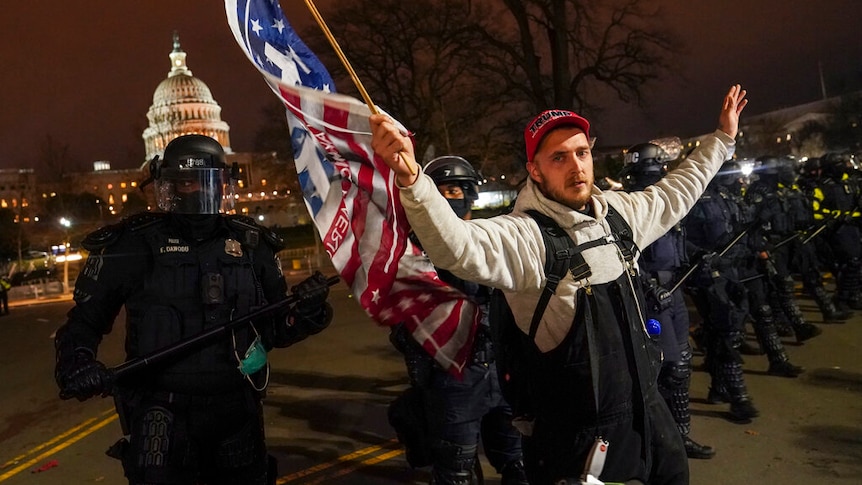 A man wearing a Trump cap and waving a flag is moved along by heavily armed police.