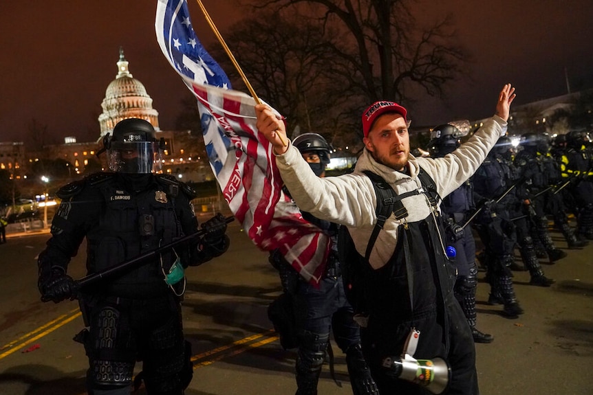 A man wearing a Trump cap and waving a flag is moved along by heavily armed police.