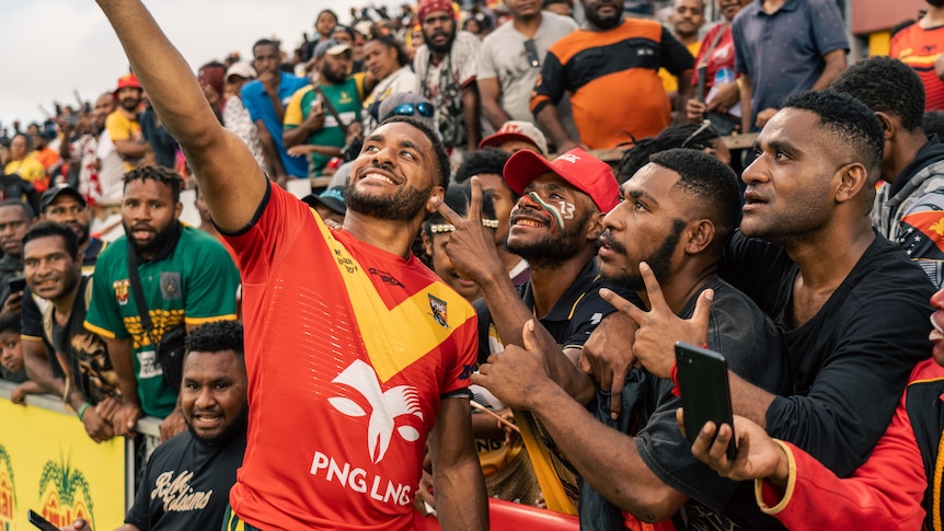 A rugby player poses for a selfie with members of the crowd, they all smile.