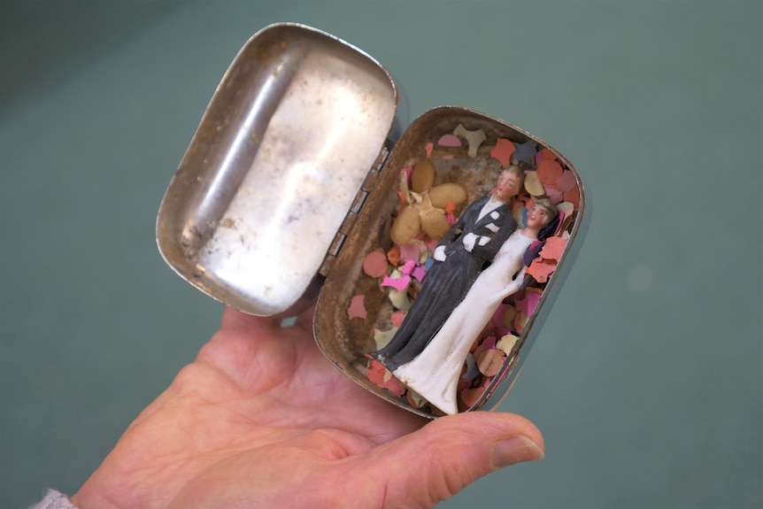 A close up of a hand holding a small tin with vintage multi-coloured confetti and a tiny plastic ornament of a bride and groom.