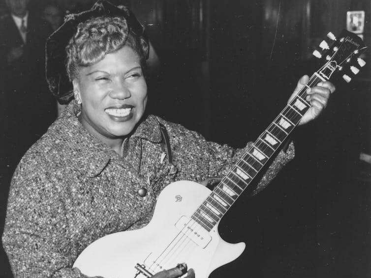 A 1957 photo of  Sister Rosetta Tharpe sitting down playing the guitar in a lounge at London Airport