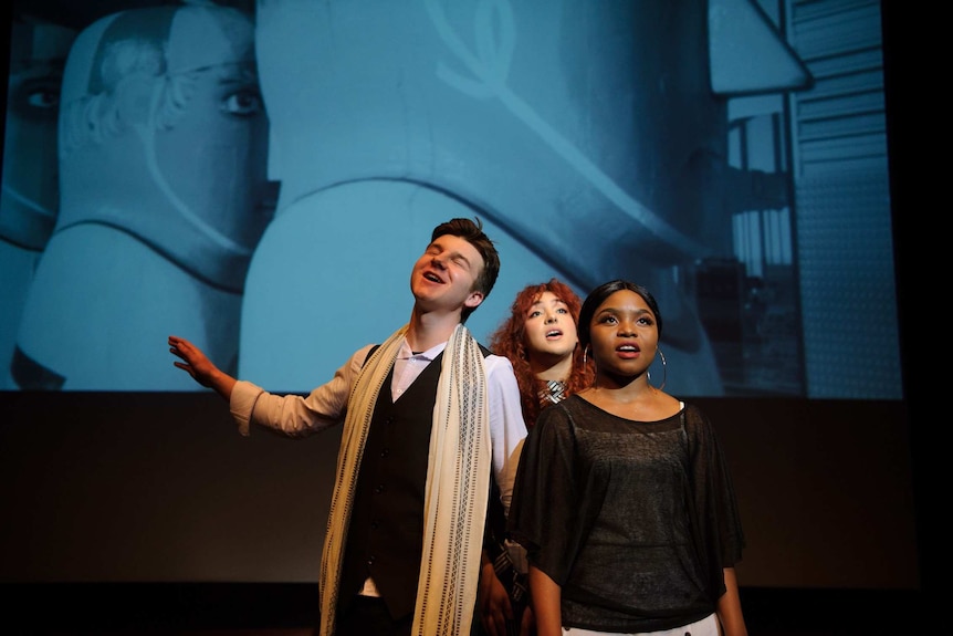 On stage, a man in formal clothes tips his head back and smiles, two women sing next to him