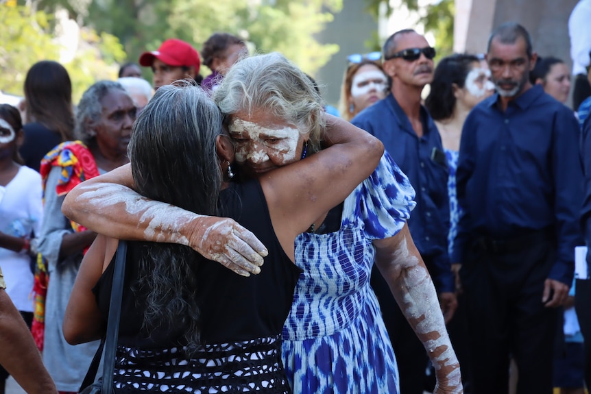 Two women embracing outdoors, with a crowd of people in the background.