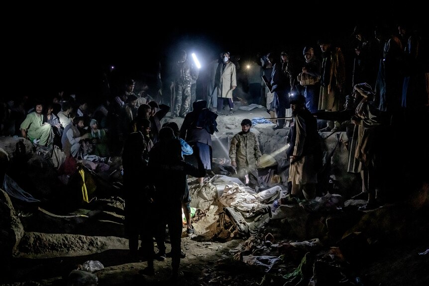 A large group of Taliban members holding electric torches stand around a hole in the ground filled with garbage at night.