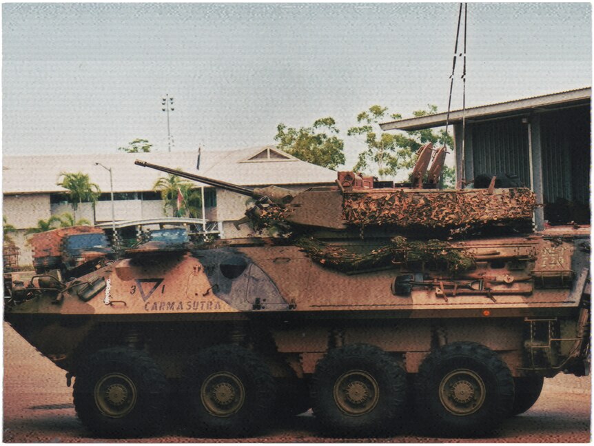 An eight-wheeled light armoured vehicle sits parked. Behind it are some low buildings and trees.