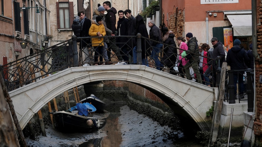 About a dozen people dressed for cold weather climb the stairs over an old bridge above a muddy ditch with two boats in it.