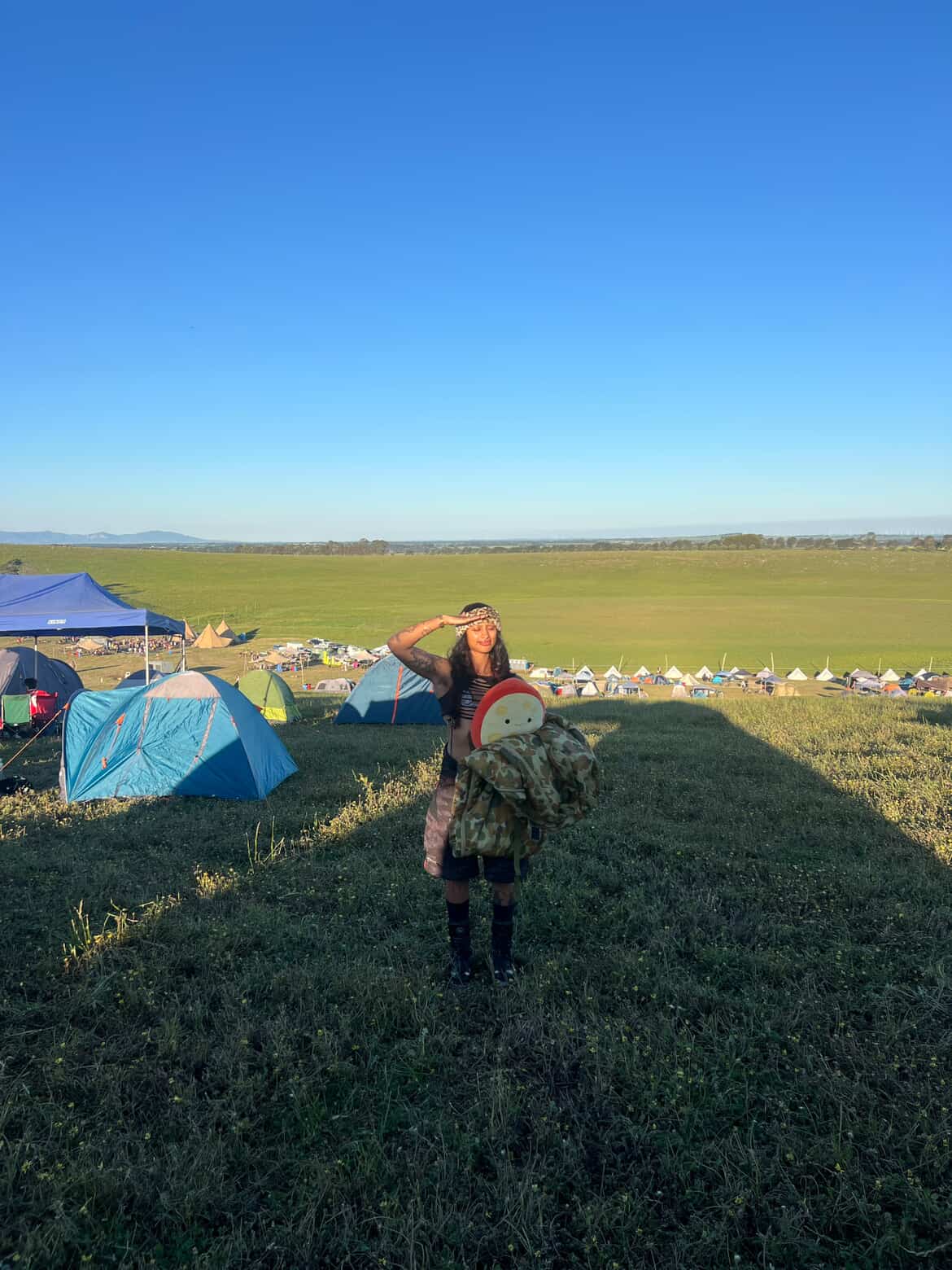 Uma mulher em um festival de música fica em frente às tendas. Ela protege o rosto do sol com a mão. 