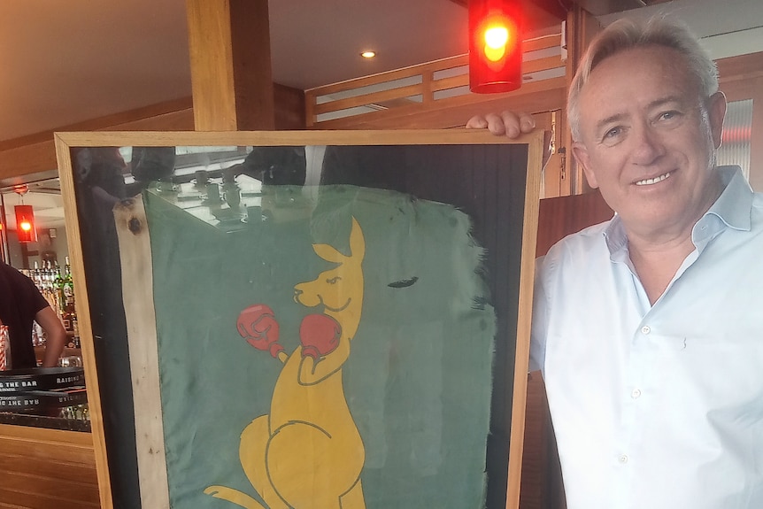 smiling holding a weathered boxing kangaroo flag in a frame.