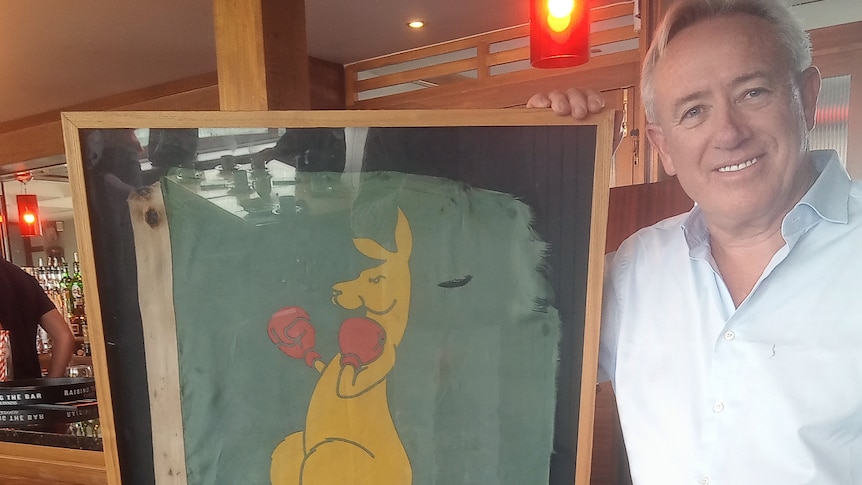 man smiling holding a weathered boxing kangaroo flag in a frame.