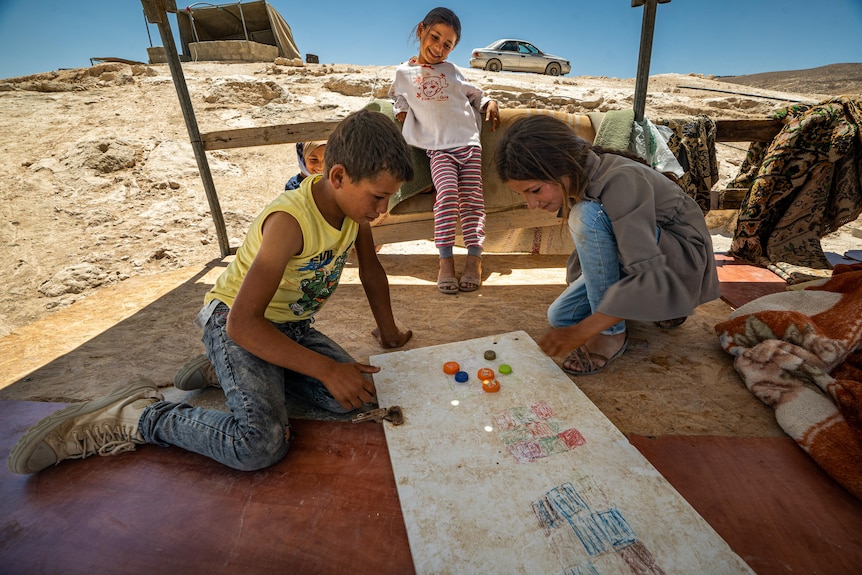 Two children play a board game together while a third watches on. They are all on a raised platform in a dusty, rocky area