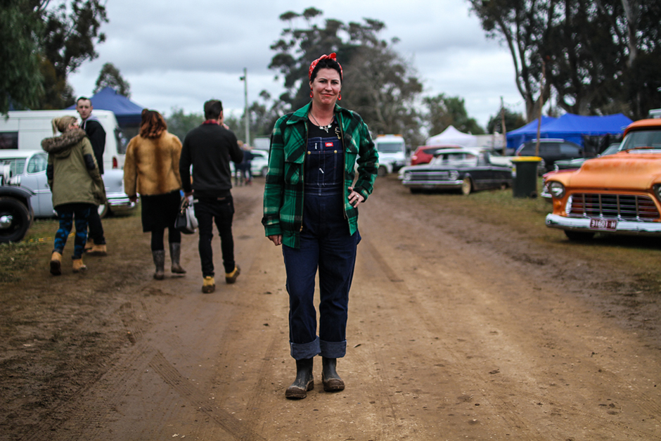 A woman in a green flannel jacket and red bandanna stands on a dirt track with vintage cars in the background.