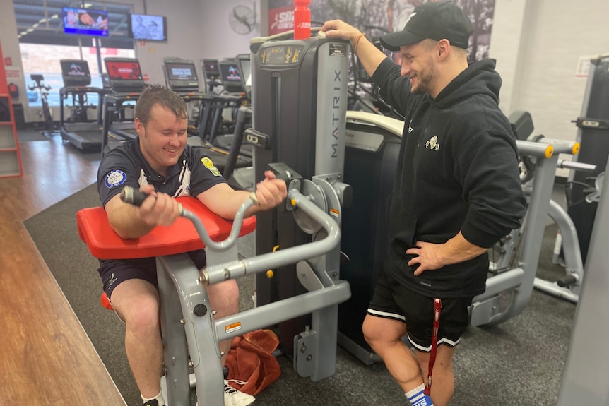 Zac on gym equipment for arm strength and Liam standing near him, inside a gym. 