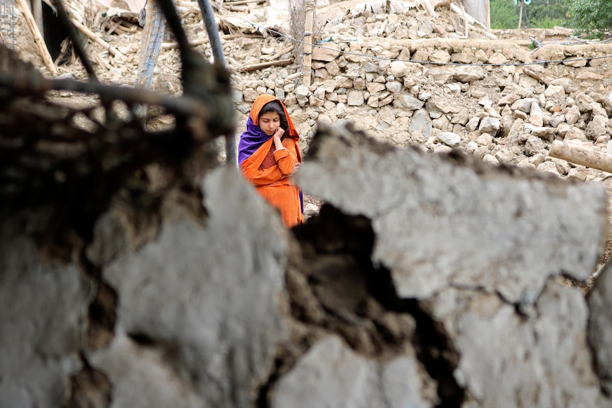 A girl in an orange outfit and headscarf pictured in distance with rubble in foreground.