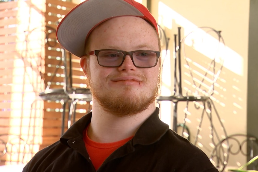 A young man with glasses, an orange beard and red cap smiles at the camera