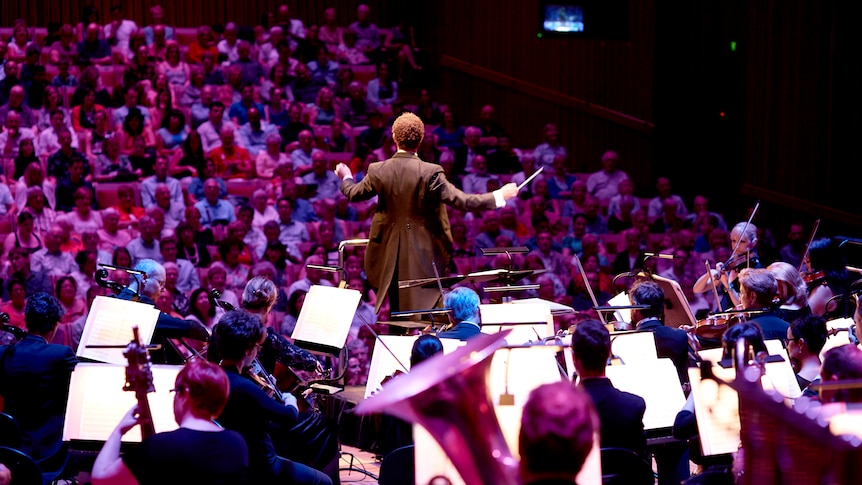 A view from behind the orchestra of the audience a the Sydney Opera House and a conductor's back as he conducts the audience