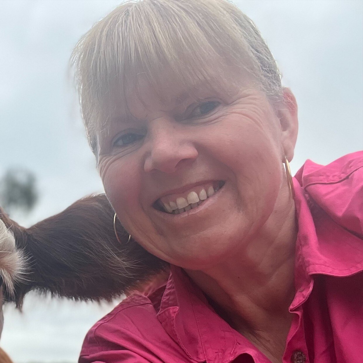 A close up shot of a woman with blonde hair, big hoop earrings and a pink shirt smiling.