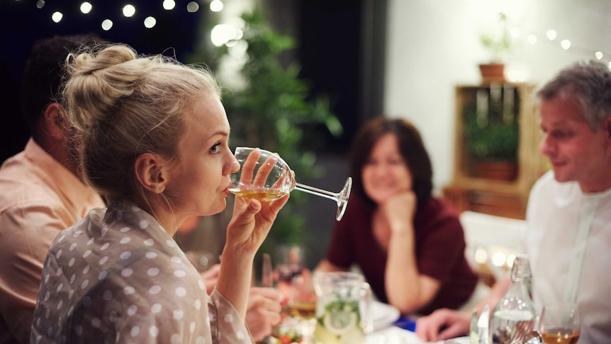 A woman holds a wine glass to her mouth and raises an eyebrow, while a blurred group of people sit at a dinner table with her.