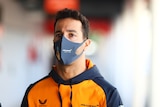 F1 driver Daniel Ricciardo stands in front of the camera in a black and orange team top while wearing a mask.