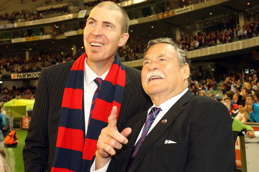 Jim Stynes and Ron Barassi smile while wearing suits inside the MCG