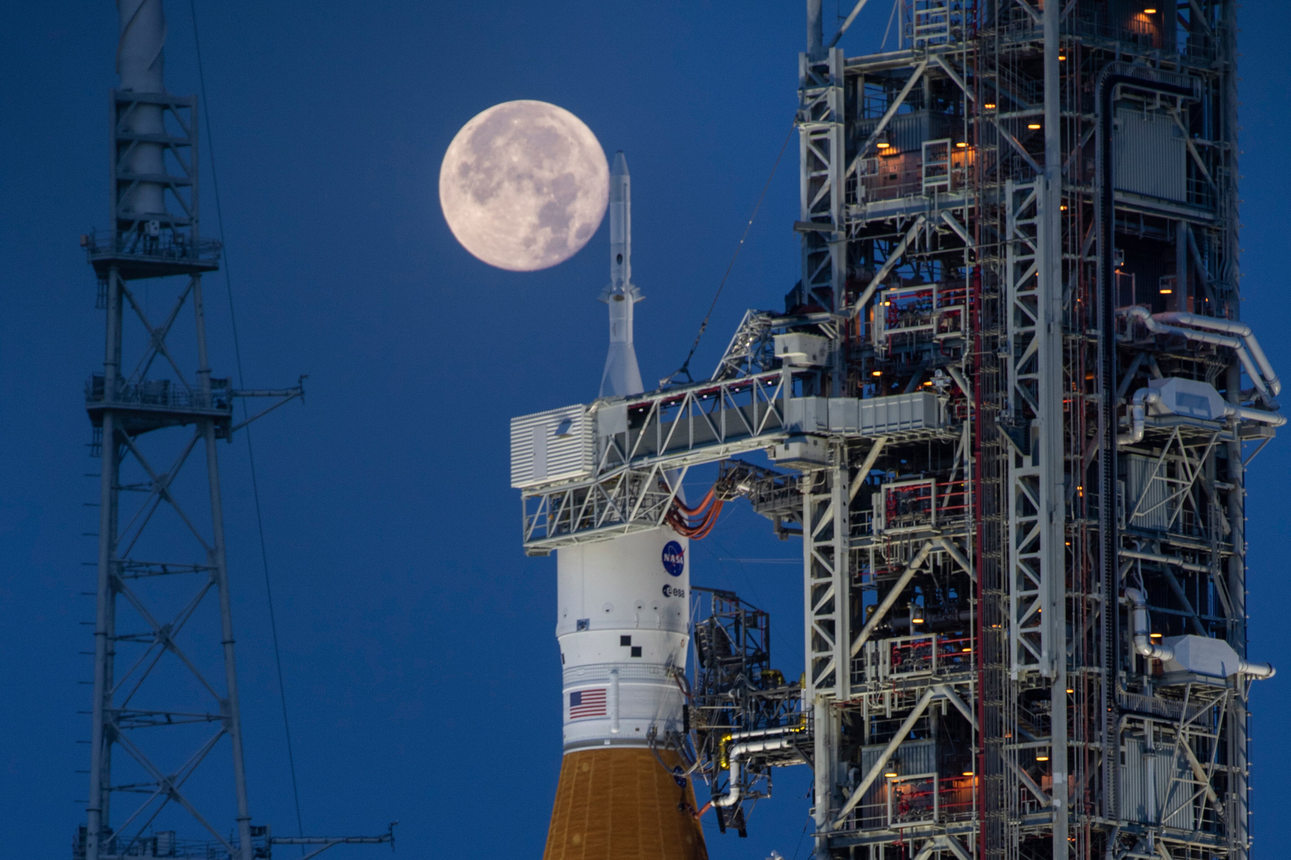 A full Moon illuminates the sky behind a rocket with a US flag and NASA logo on it, which sits on a launchpad
