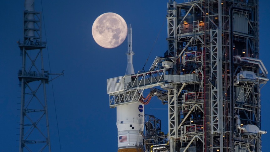A full Moon illuminates the sky behind a rocket with a US flag and NASA logo on it, which sits on a launchpad
