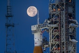 A full Moon illuminates the sky behind a rocket with a US flag and NASA logo on it, which sits on a launchpad