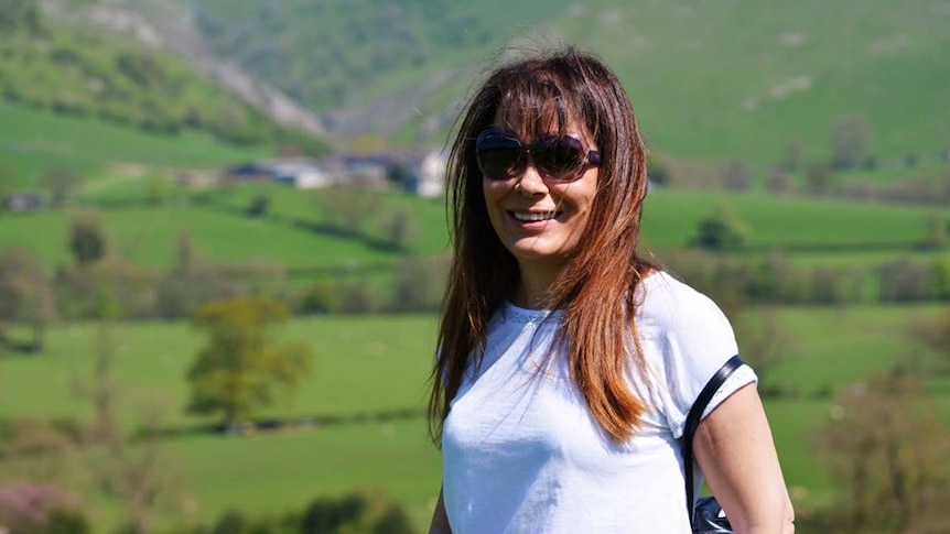 A woman with medium-length brown hair stands in front of mountains