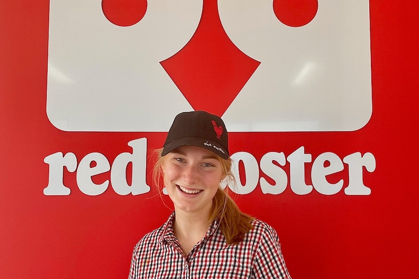 A young woman wearing a red and white checkered shirt and black Red Rooster cap stands in front of a Red Rooster sign.