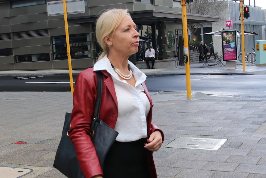 Defence counsel Justine Fisher stands on a footpath near a set of traffic lights wearing a red jacket and white shirt.