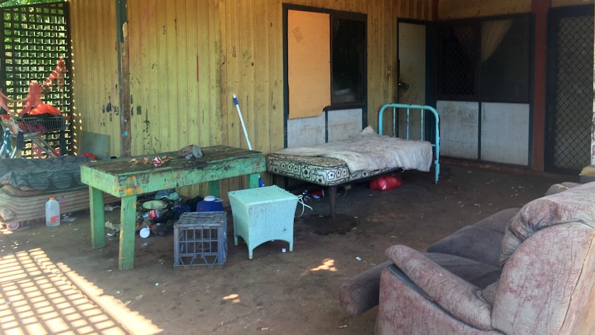 A dirty, dilapidated house with a bed and sofa outside under the porch.