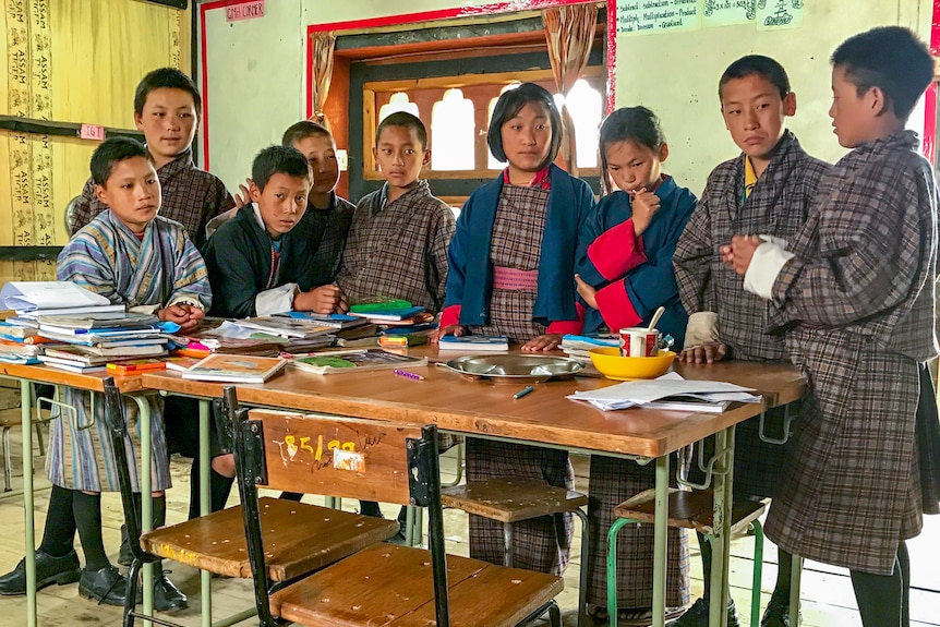 Primary school students in Bhutan stand around a table in a classroom, wearing their national dress.