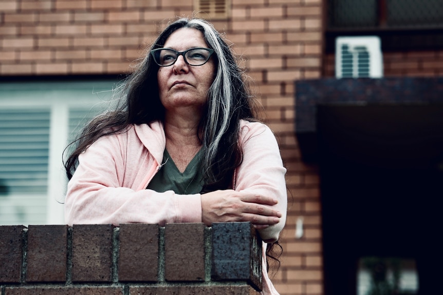 A woman with dark long hair and glasses stands in front of a 1950s unit block.
