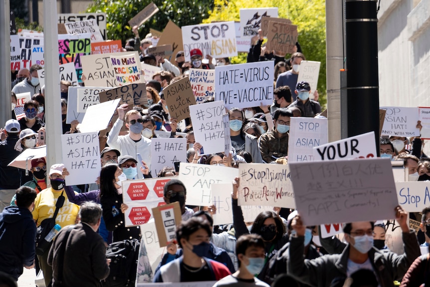 A large crowd of protesters wear masks and carry signs as they walk on sunny street.